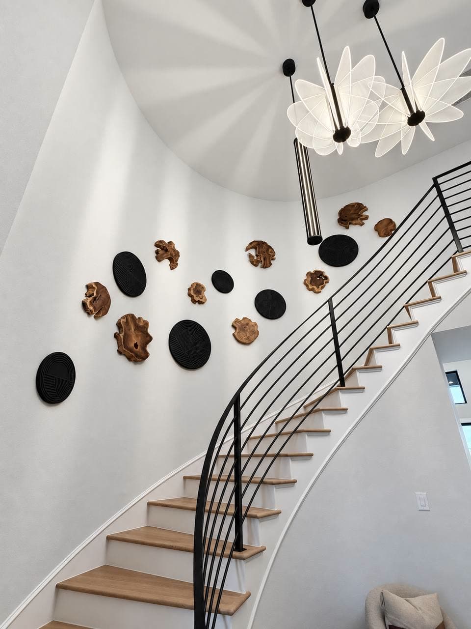 Curved stairwell wall hung with black ribbed discs and carved wood objects, beside a black rail and a petal chandelier.