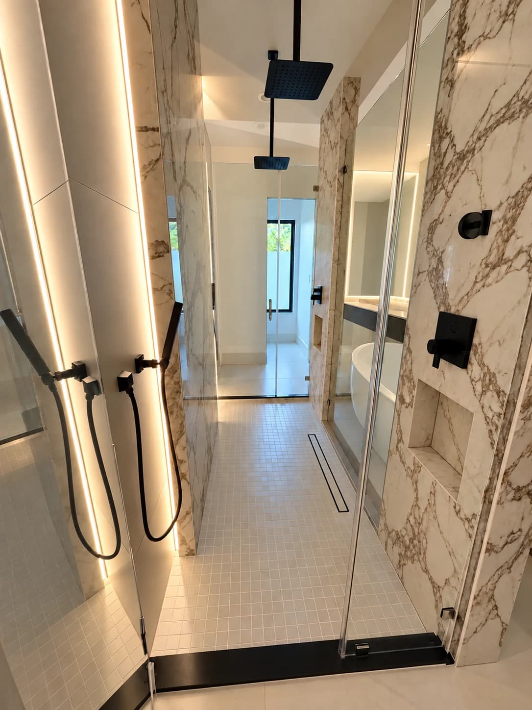 Long walk-through shower with gold-veined marble walls, two black ceiling-mounted rain heads, black handshowers, recessed niches, a linear drain and a white square mosaic floor.