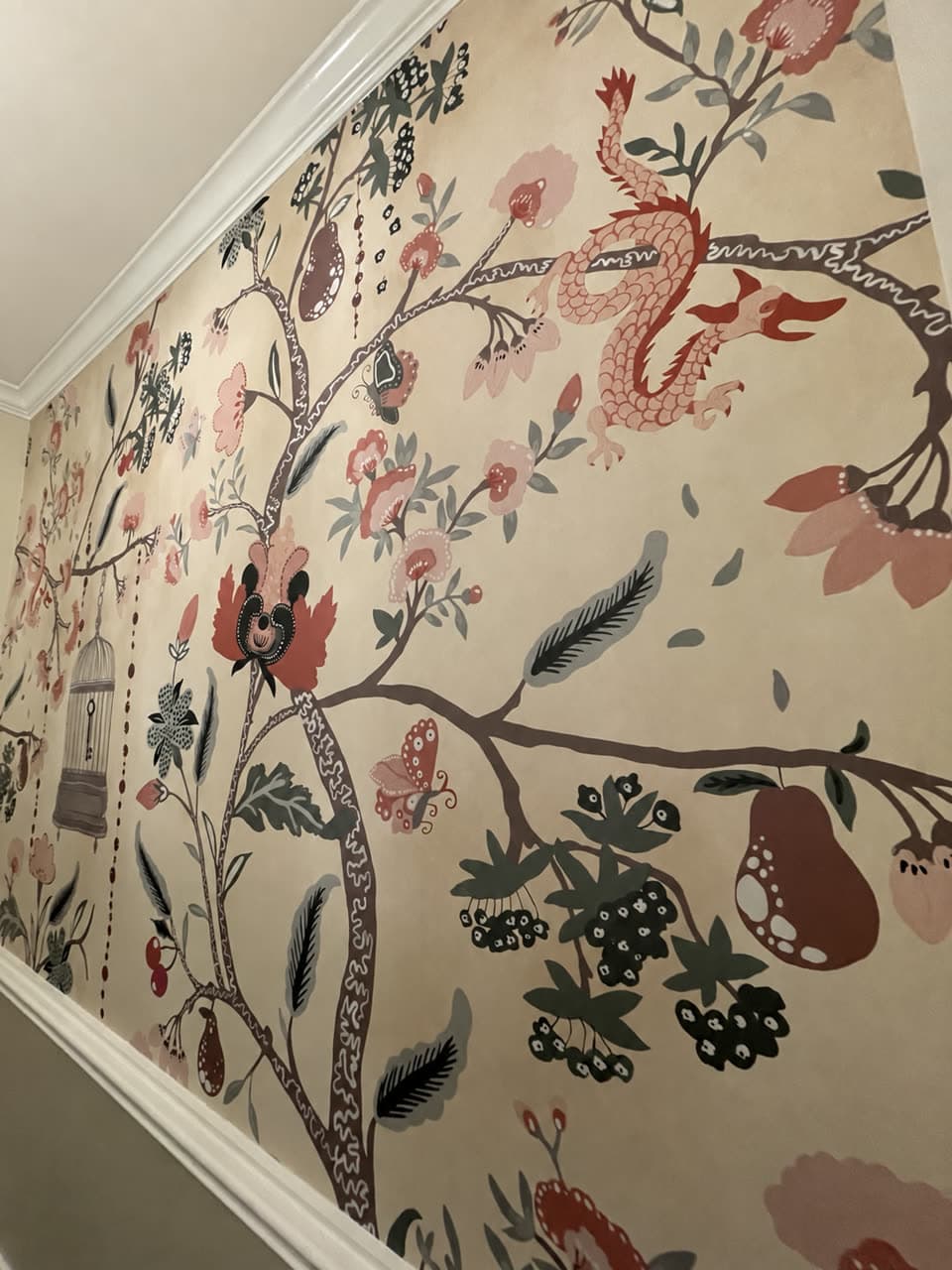Stairwell wall in a cream botanical mural — branches, fruit, birds and a red dragon — above white chair rail and below crown moulding.