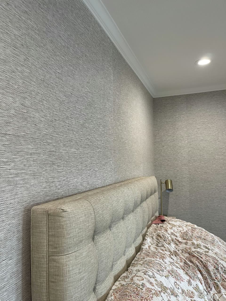 Bedroom corner in fine grey and cream grasscloth wallpaper meeting white crown moulding above an upholstered headboard.