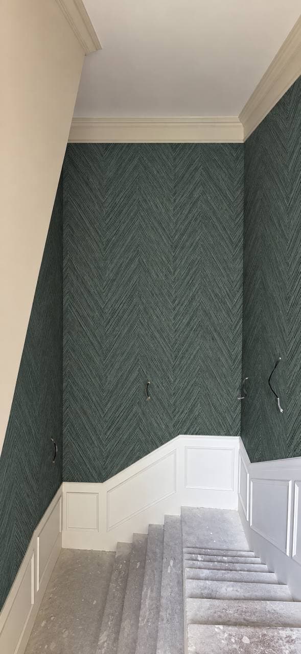 Stairwell wall in dark green herringbone grasscloth above white panel wainscoting, mid-renovation.