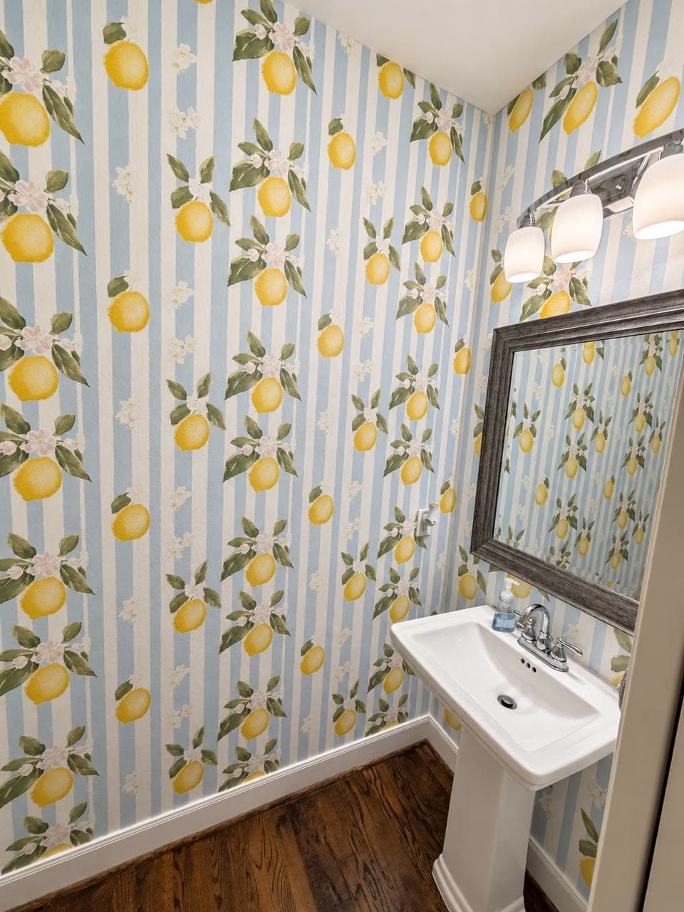 Powder room papered in a blue and white stripe with lemons and blossom, above a pedestal sink and a dark framed mirror.