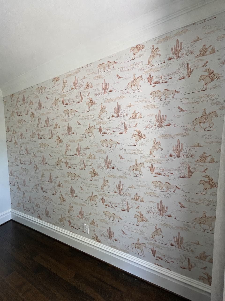 Accent wall in a rust-on-cream toile of riders, horses and cacti, meeting white baseboard and a dark wood floor.